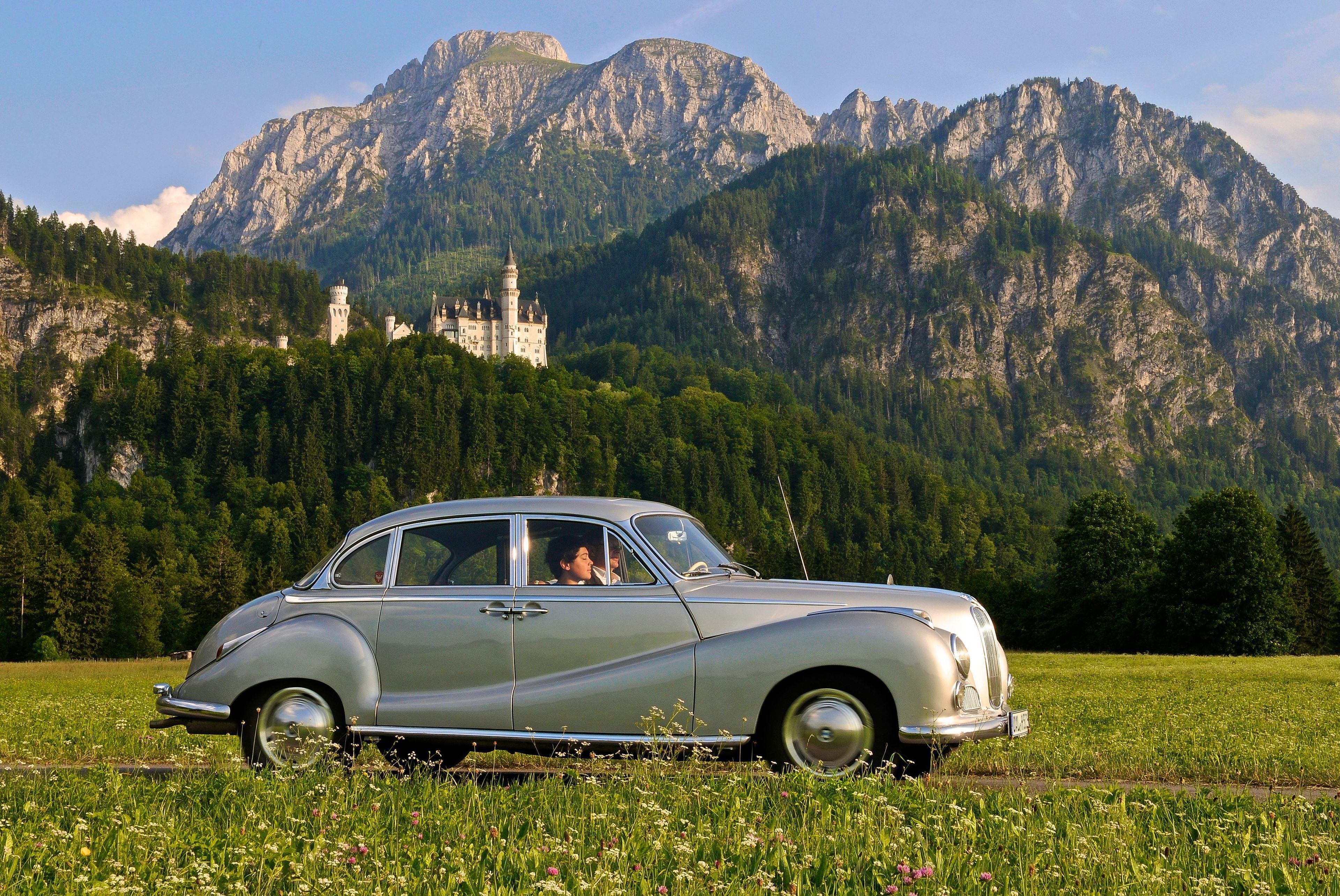 A vintage car parked in a field with a castle and mountains in the background.