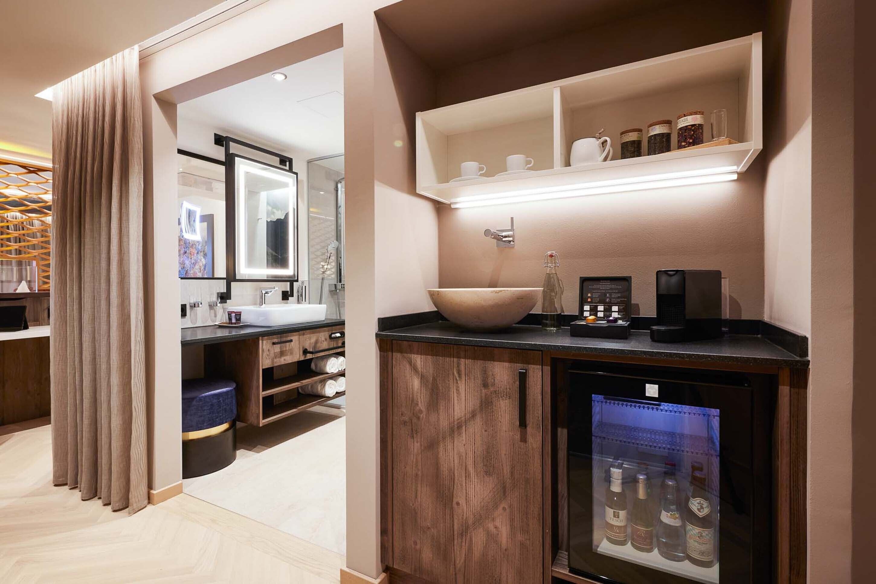 A modern bathroom with a large mirror, a sink, a vanity, a coffee machine, and a refrigerator with beverages.