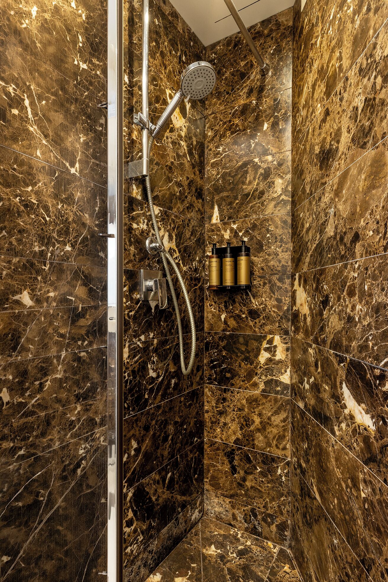 A marble bathroom with a shower, a glass door, and a shelf with toiletries.