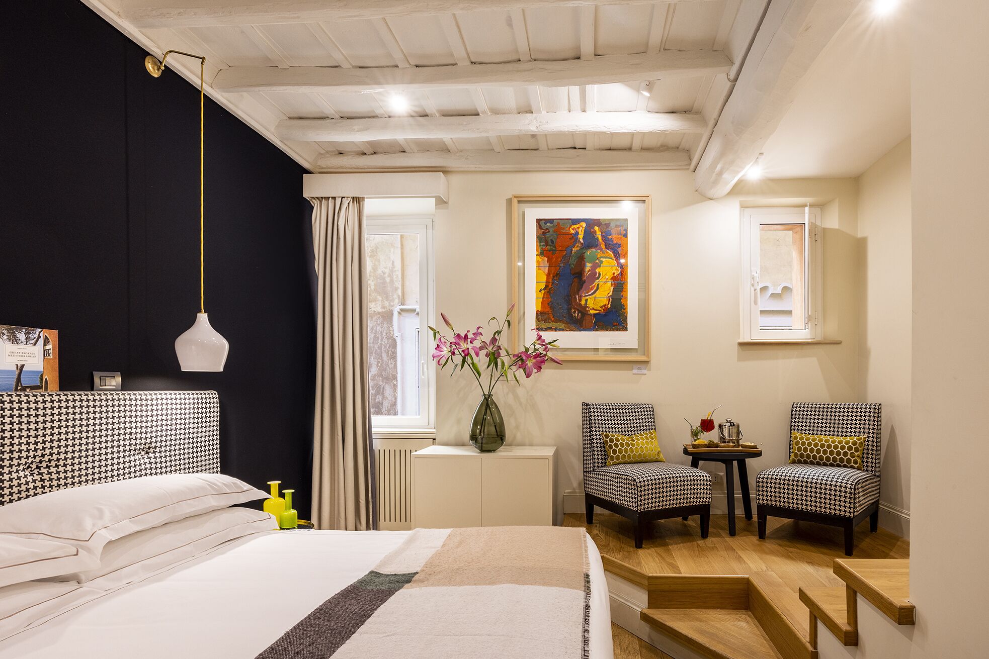 Habitación de hotel con cama, dos sillas, una mesita, un florero con flores, un cuadro en la pared y una ventana con cortinas.