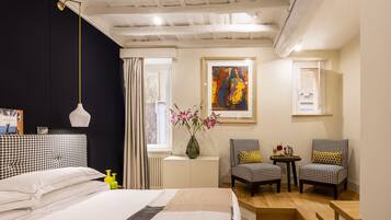 Habitación de hotel con cama, dos sillas, una mesita, un florero con flores, un cuadro en la pared y una ventana con cortinas.