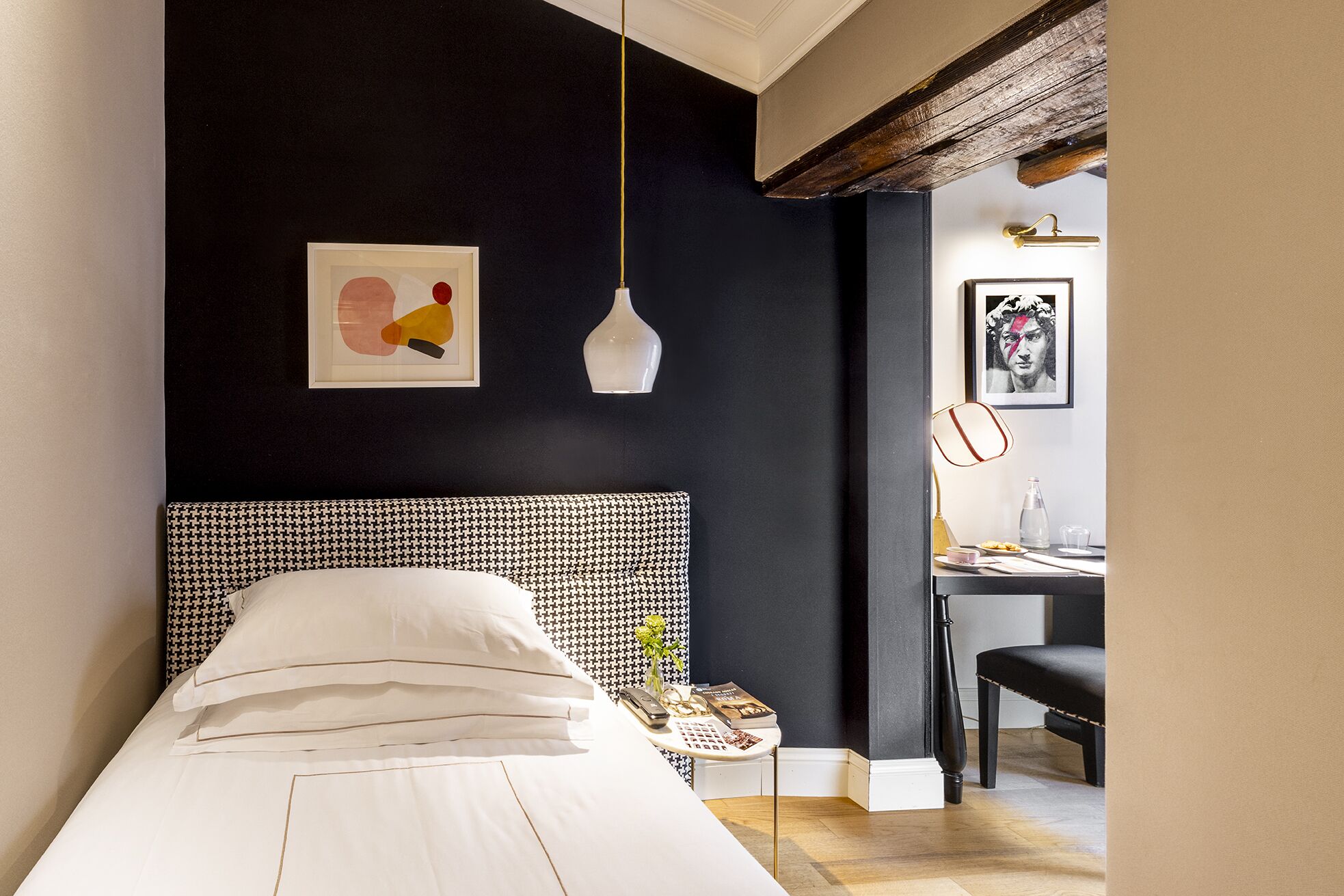 Ein Schlafzimmer mit schwarzer Wand, einem weißen Bett, einem kleinen Tisch mit Büchern und einer Vase sowie einem Schreibtisch mit Lampe und gerahmtem Bild.