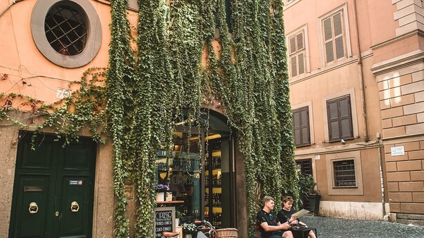 A cobblestone street with a building covered in ivy, a bicycle, and a person sitting on a chair. - Nerva Boutique Hotel (Rome)