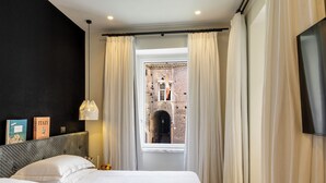 A bedroom with a bed, curtains, a TV, a wall-mounted light, and a view of an outdoor area. - Nerva Boutique Hotel (Rome)