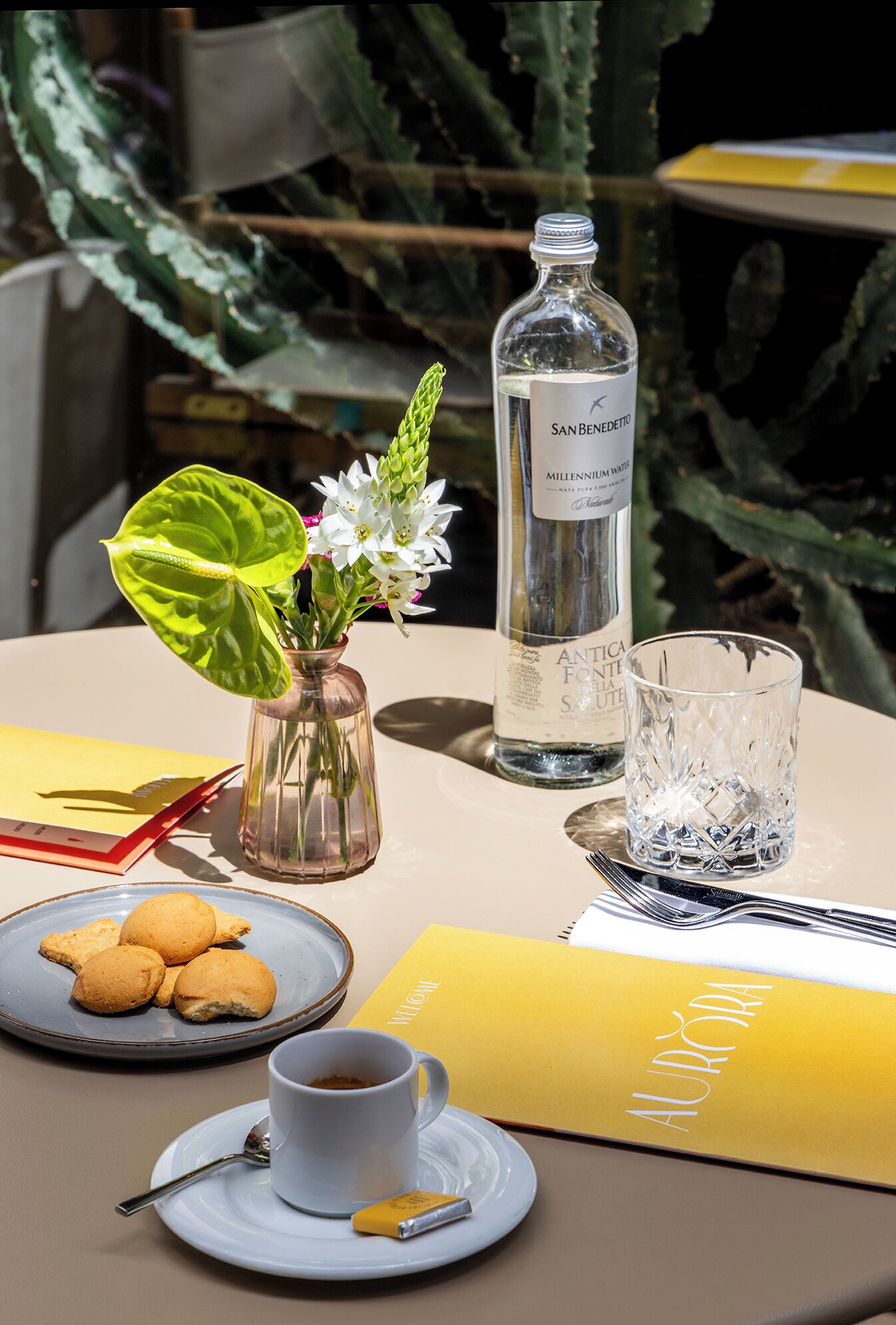 A table with a bottle of water, a glass, a cup, a plate with cookies, a spoon, a napkin, and a book.
