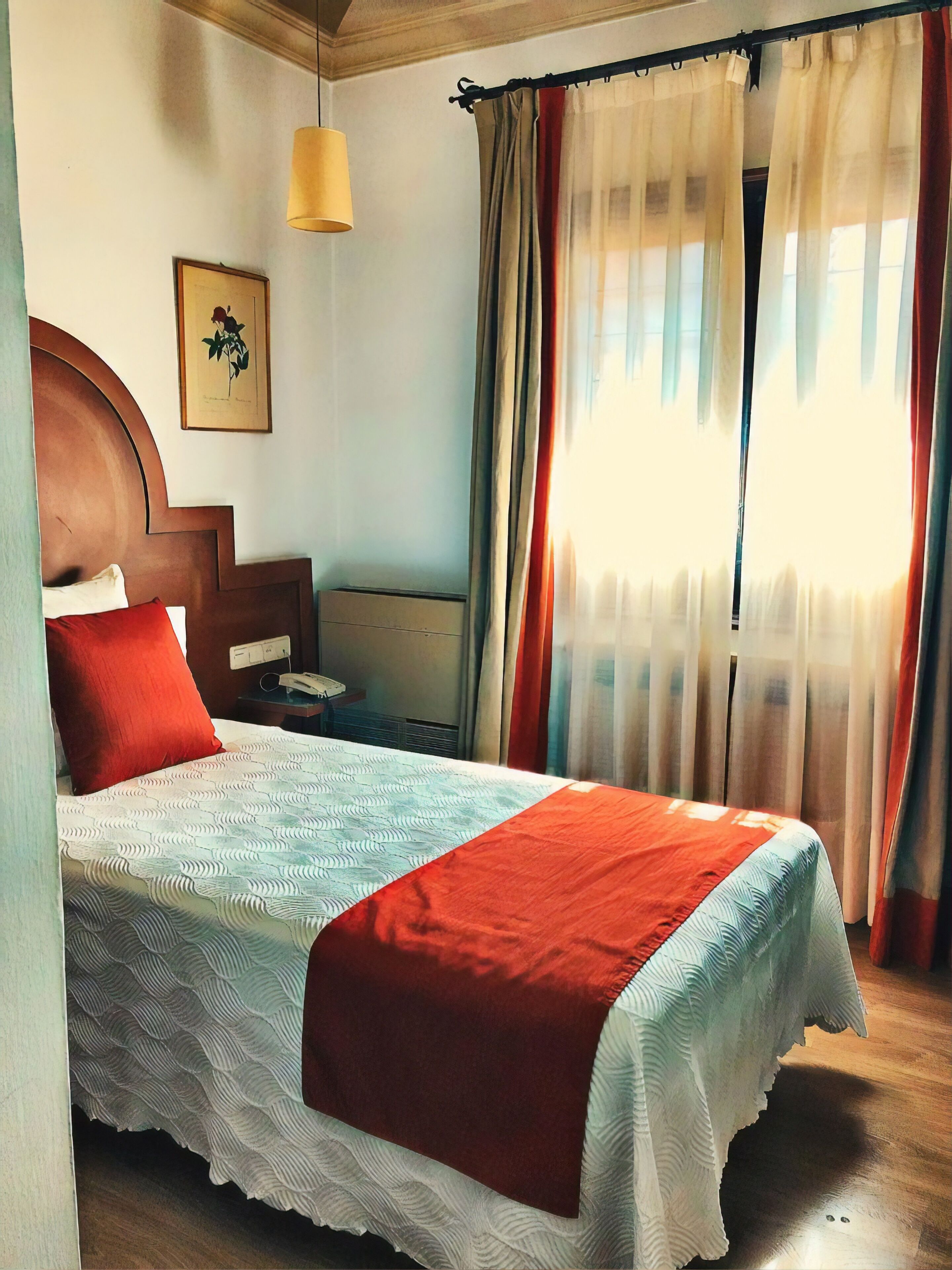 A neatly made bed with a patterned bedspread and a red accent, a wooden headboard, a bedside table with a lamp, and a wall-mounted picture.