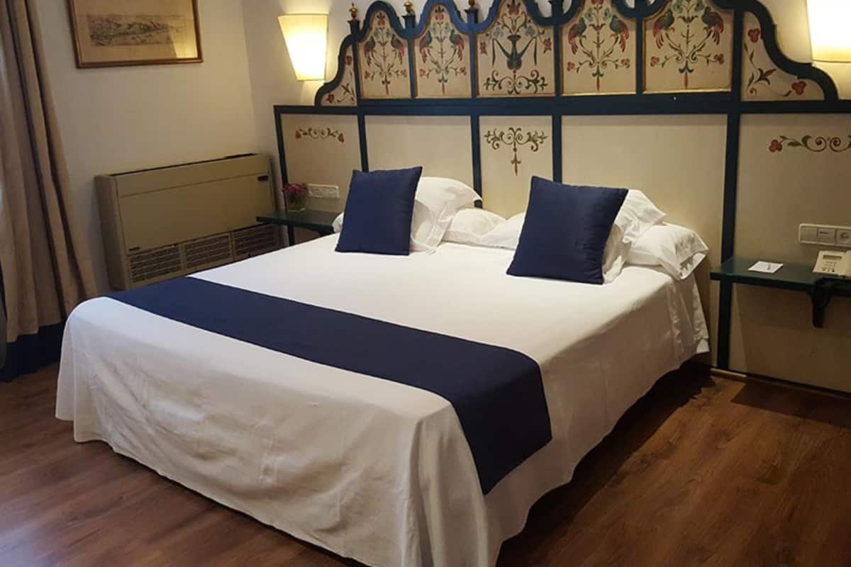 Uma cama bem arrumada com colcha branca e azul marinho, ladeada por duas mesas de cabeceira com abajures, e um painel decorativo com motivos florais.