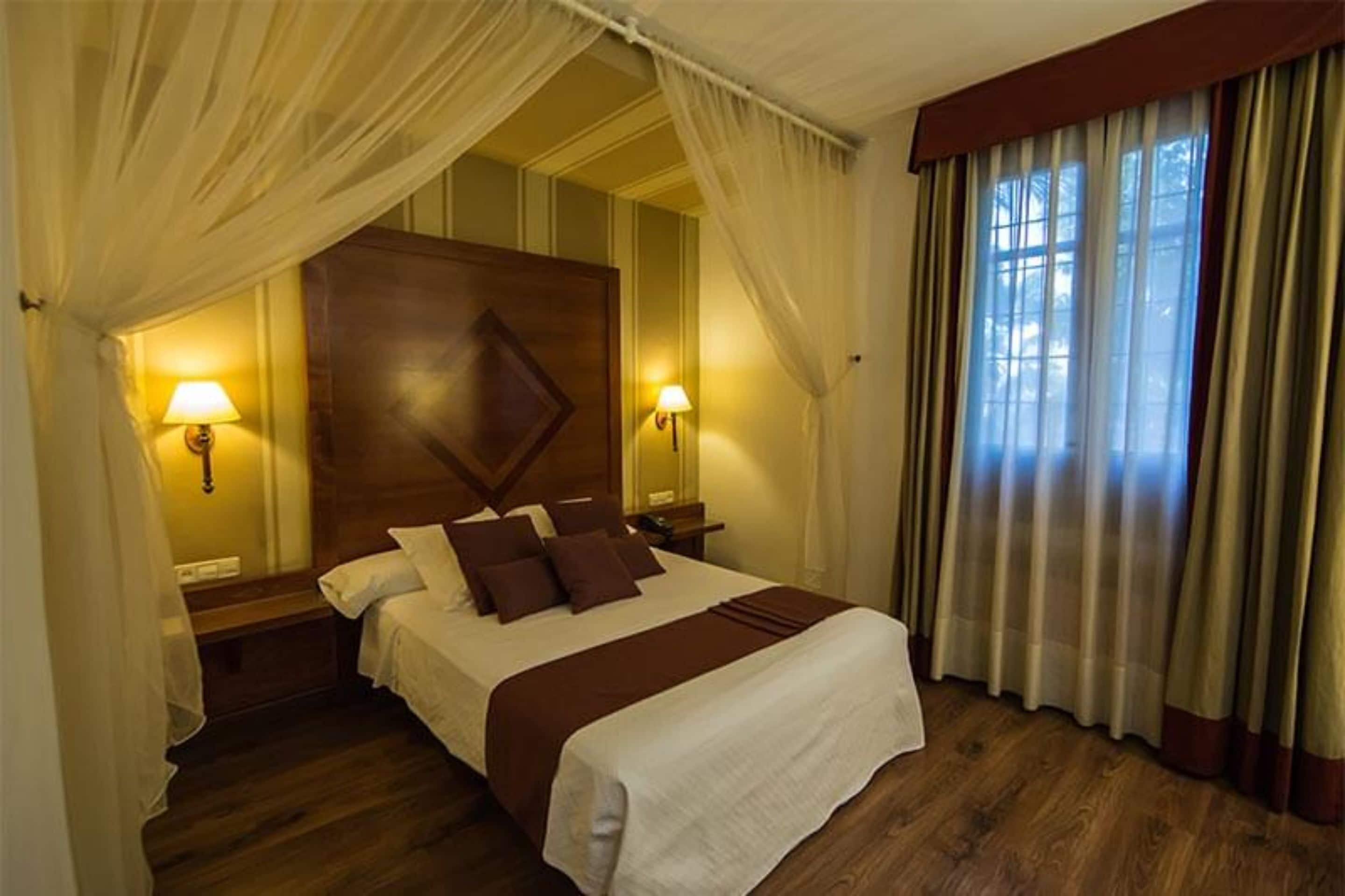Una habitación de hotel con una cama grande, dos lámparas de noche, un cabecero de madera y cortinas de encaje.