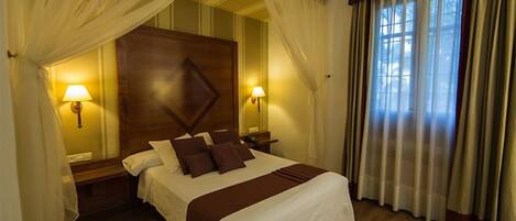 A hotel room with a large bed, two bedside lamps, a wooden headboard, and sheer curtains.