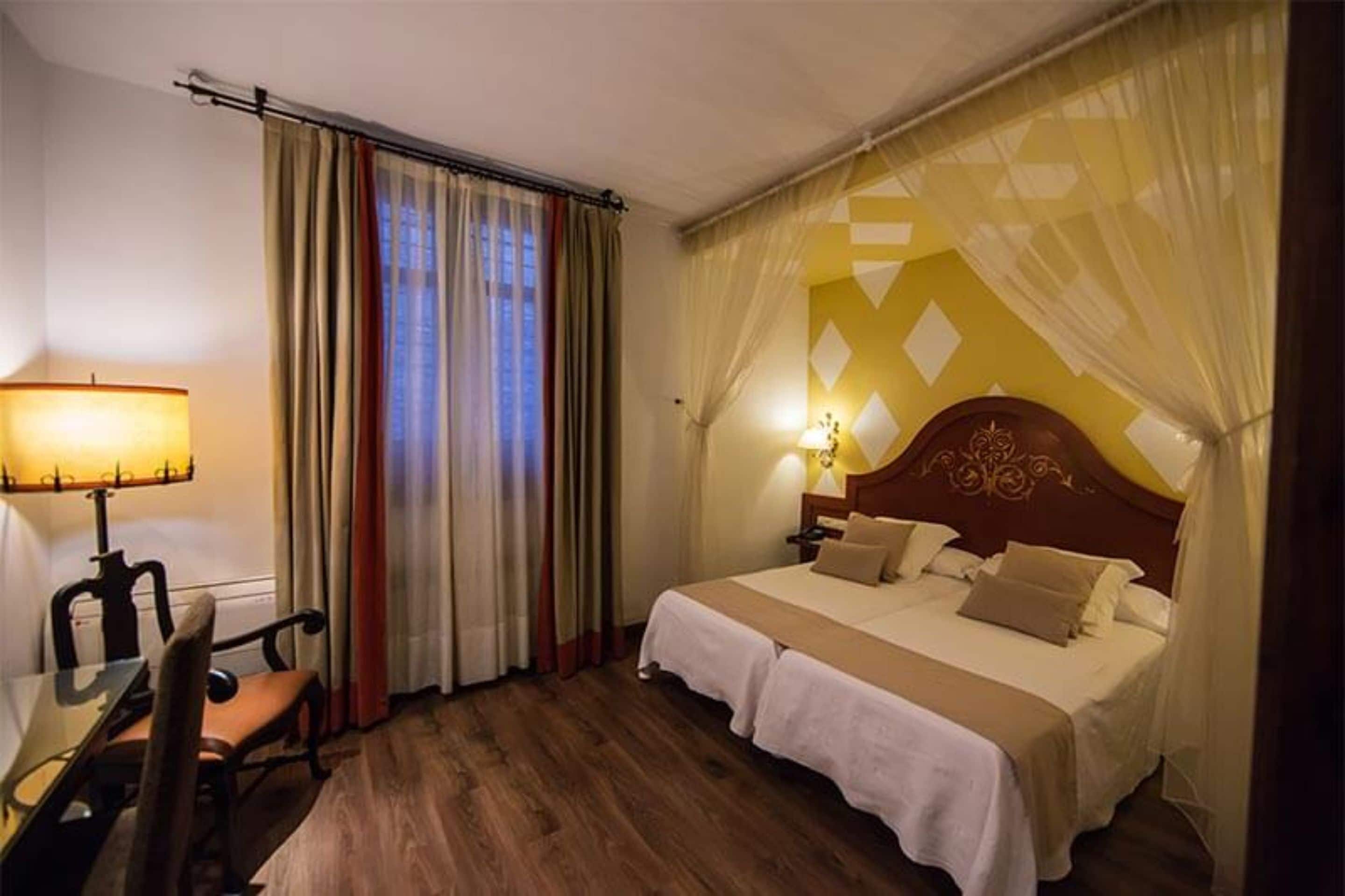 Hotelkamer met houten vloer, een hemelbed, nachtkastje met lamp en een stoel.