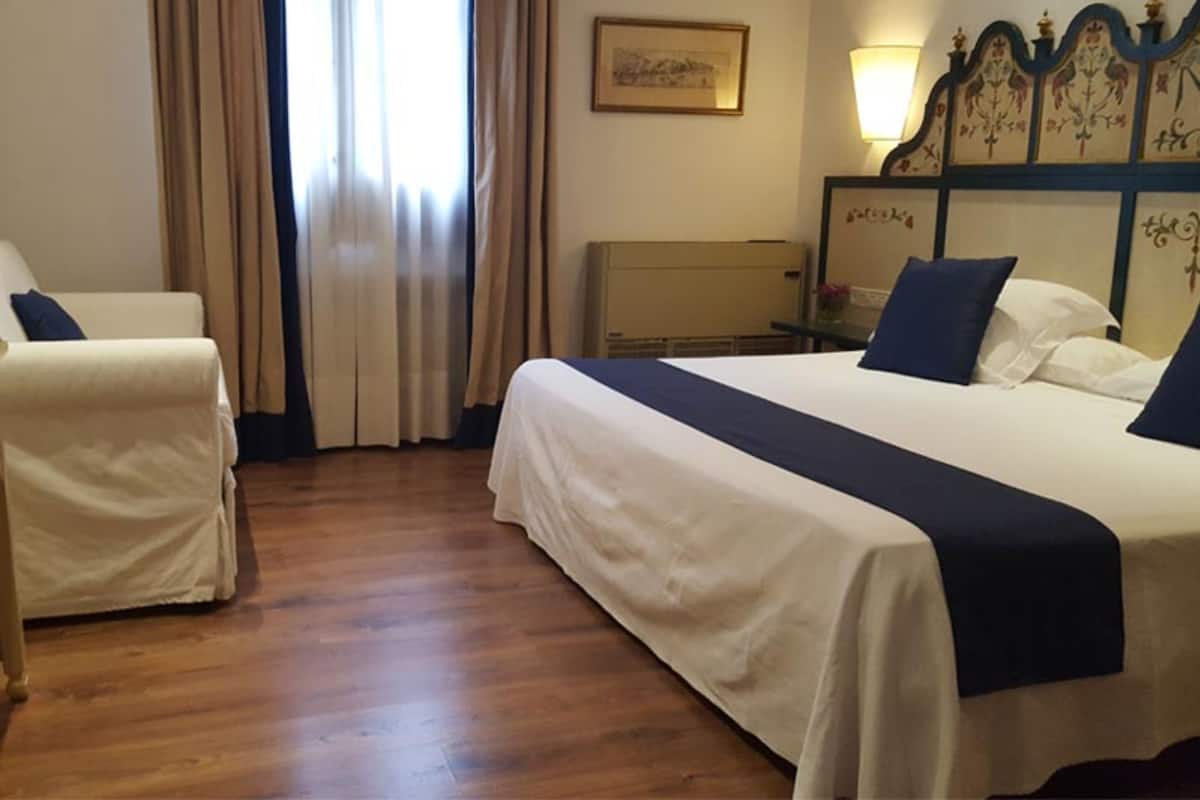 Quarto de hotel com uma cama grande, uma poltrona, uma escrivaninha e uma luminária.