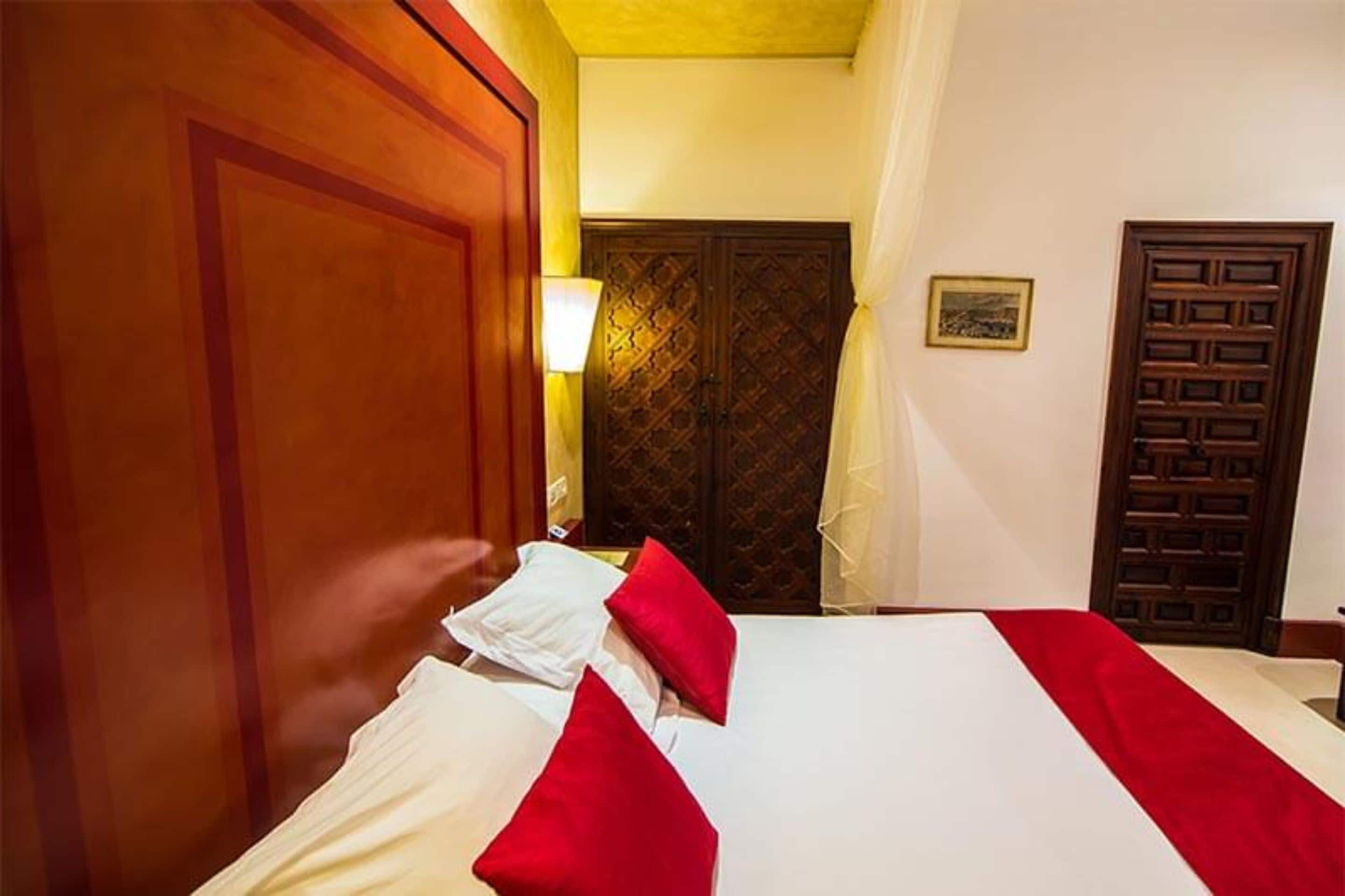 Habitación de hotel con una cama grande, dos almohadas rojas, un armario de madera y una lámpara de pared.