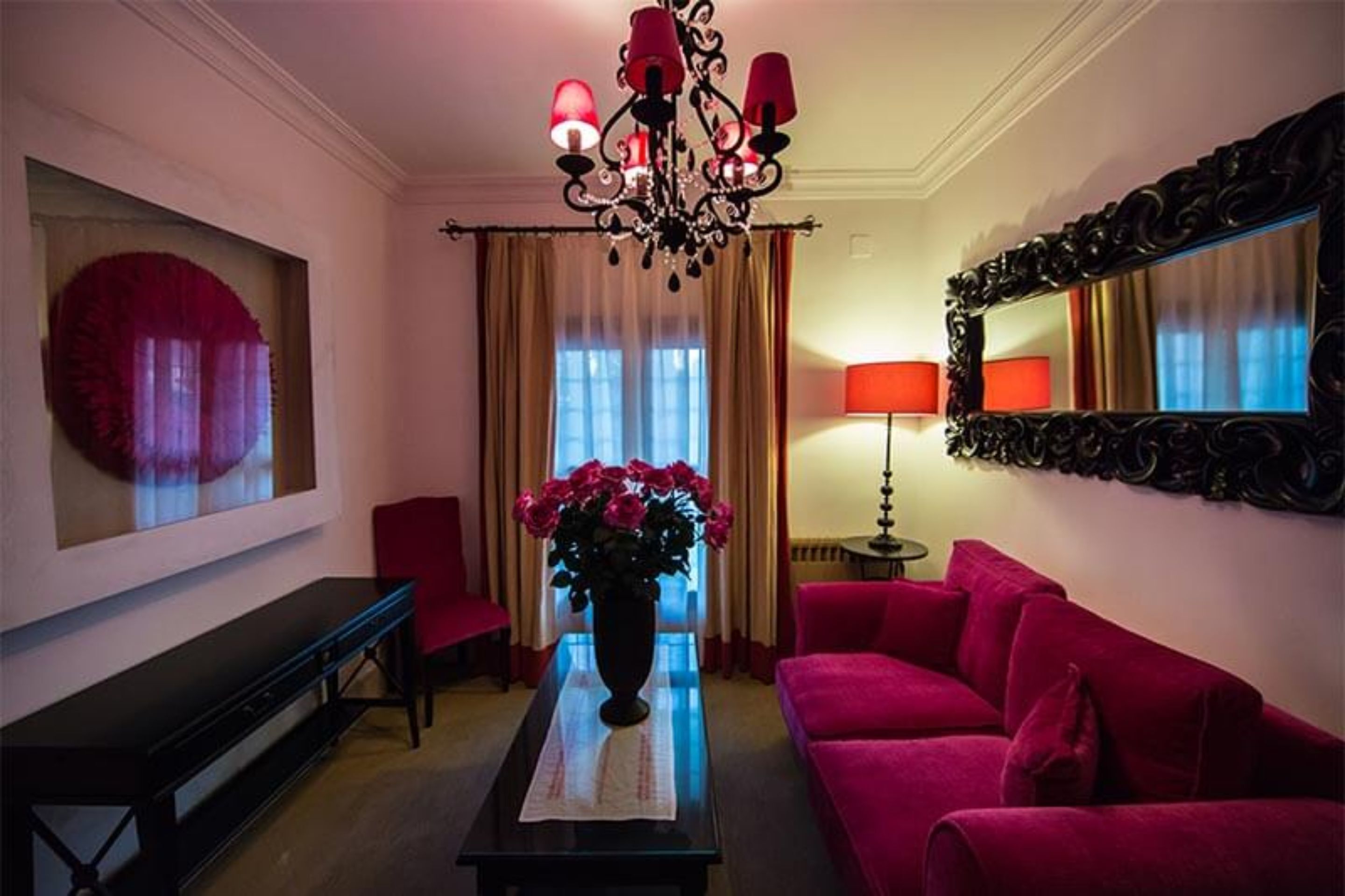 Una stanza con un divano rosso, una panca nera, un vaso di fiori, un lampadario, uno specchio e una finestra con tende.