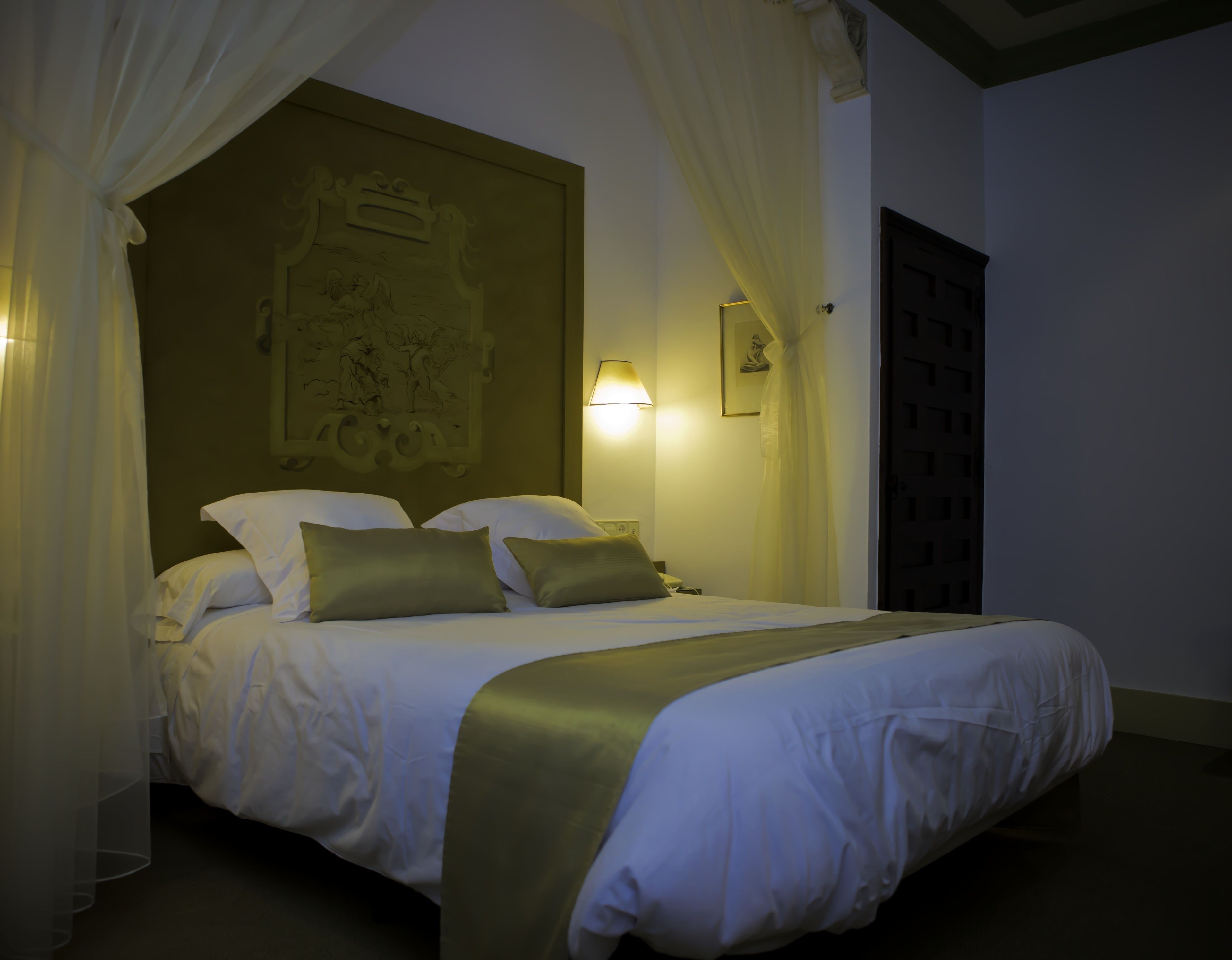 A hotel room with a bed, white bedding, a canopy, a wall-mounted artwork, and a bedside lamp.