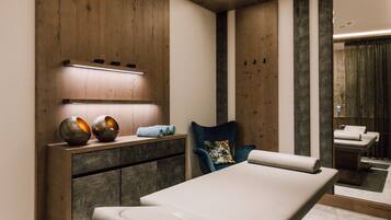 A modern spa room with a massage table, wooden panel walls, and a small seating area with a blue pillow.