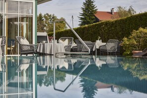 Een zwembadterras met rieten stoelen en tafels, een glazen balustrade en uitzicht op een gebouw en bomen.