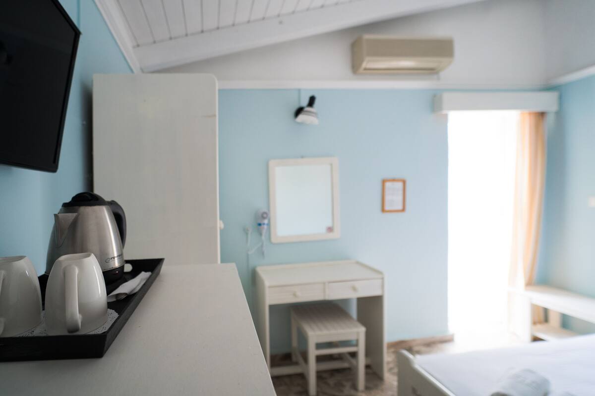 A hotel room with a desk, a small stool, a bed, a bedside table, a tray with cups and a kettle, a mirror, and a window with curtains.