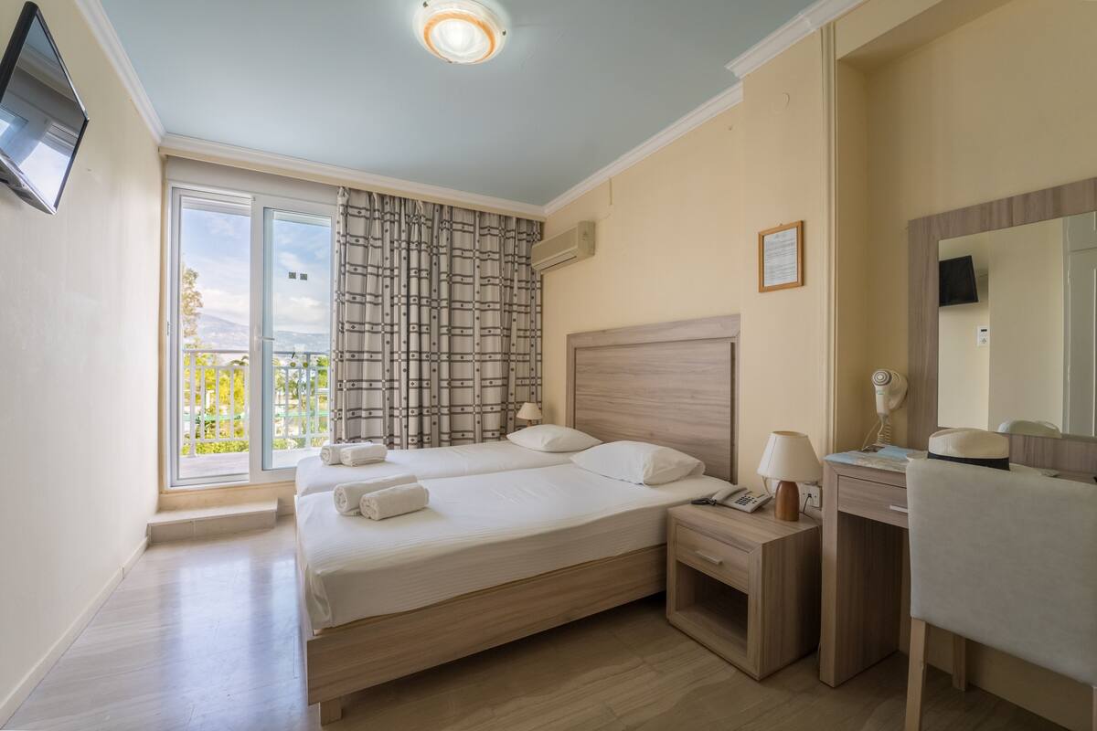 A bedroom with a large bed, a desk, a chair, a TV, and a window with a view of greenery.