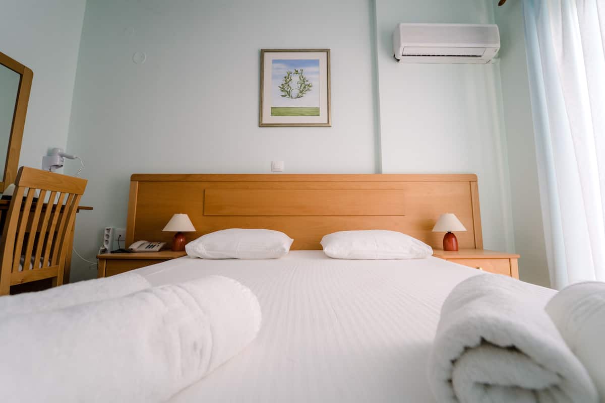 A hotel room with a wooden headboard, two bedside tables with lamps, a framed picture on the wall, and an air conditioning unit.