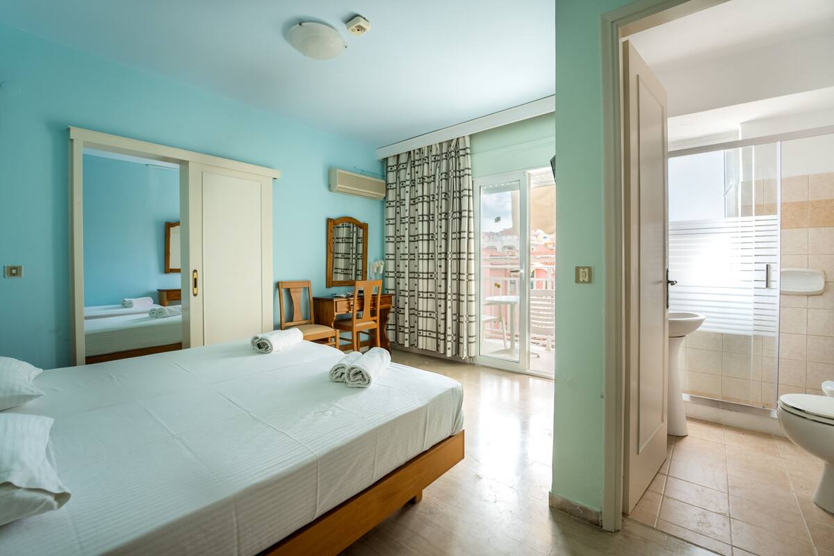 A bedroom with a bed, a desk, a chair, a mirror, and a sliding door leading to a balcony.