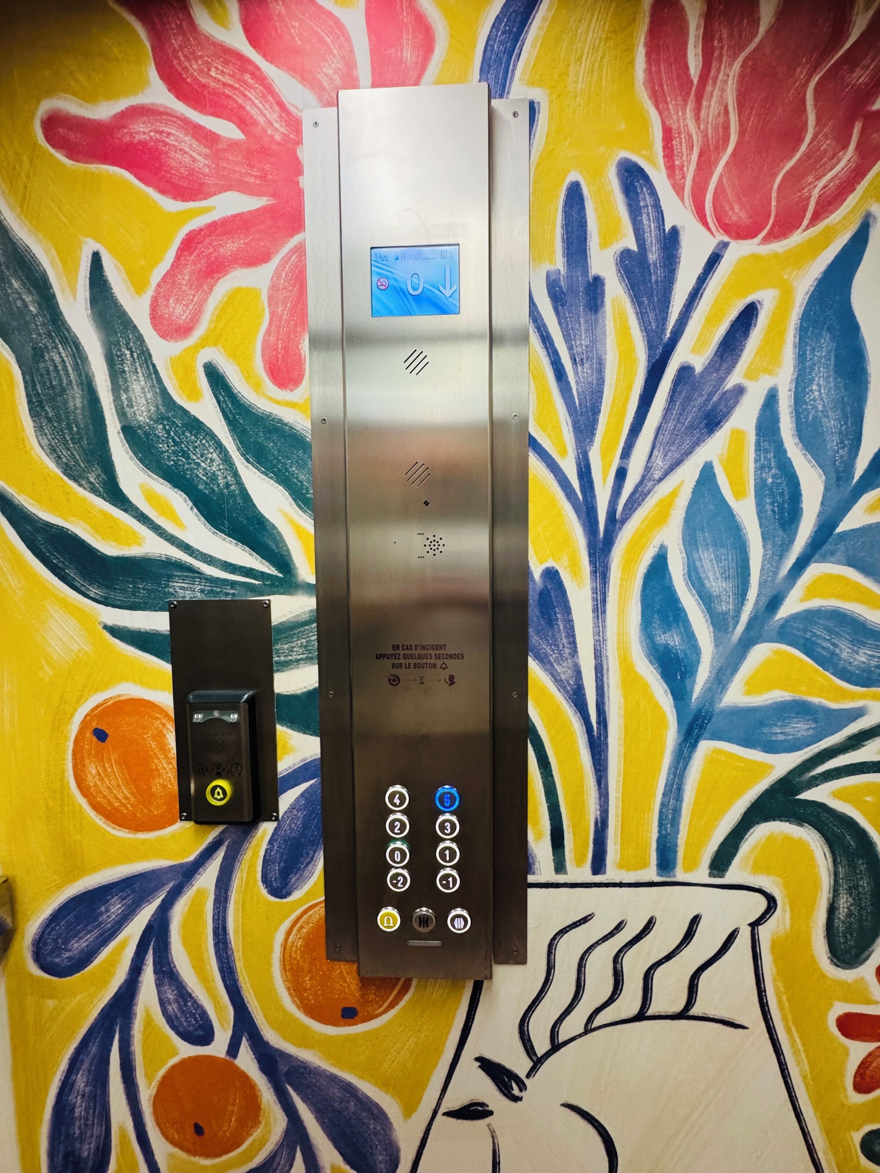 Un ascensor amb pantalla digital i teclat, davant d'un mural abstracte i colorit.