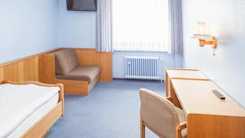 Habitación de hotel con cama, escritorio, silla, televisión, ventana con cortinas y lámpara de pared.