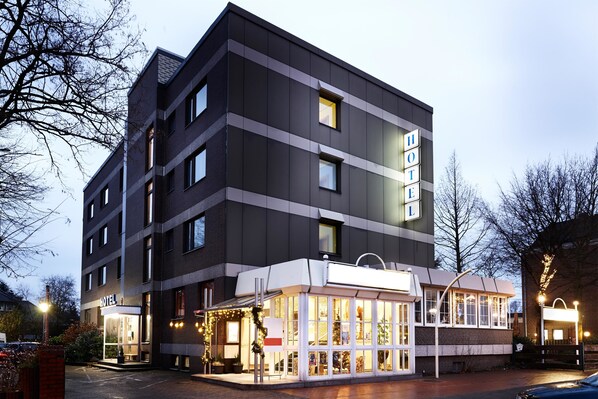 Un hotel moderno con fachada de vidrio, varios pisos y un área de restaurante visible con mesas al aire libre.