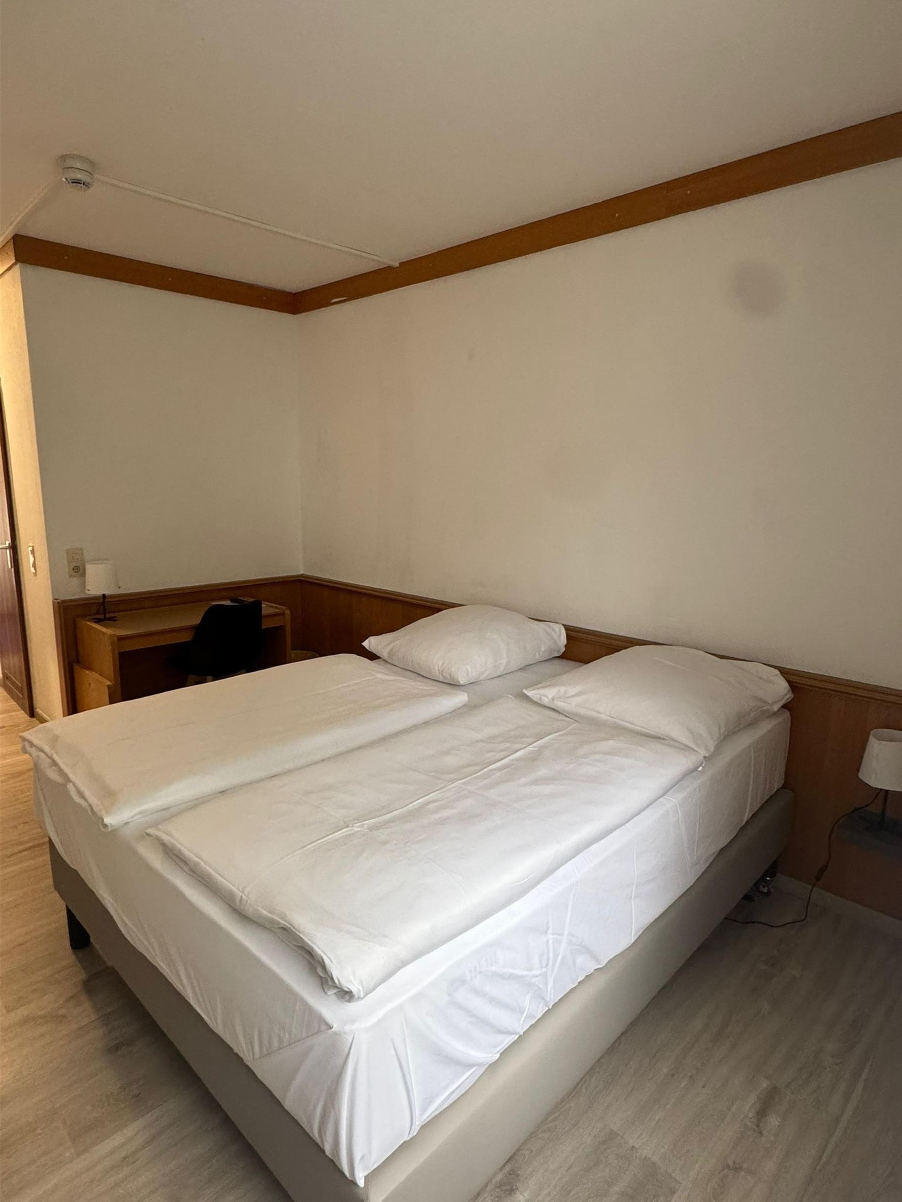 A single-bed room with a wooden headboard, a desk with a chair, and a bedside table with a lamp.