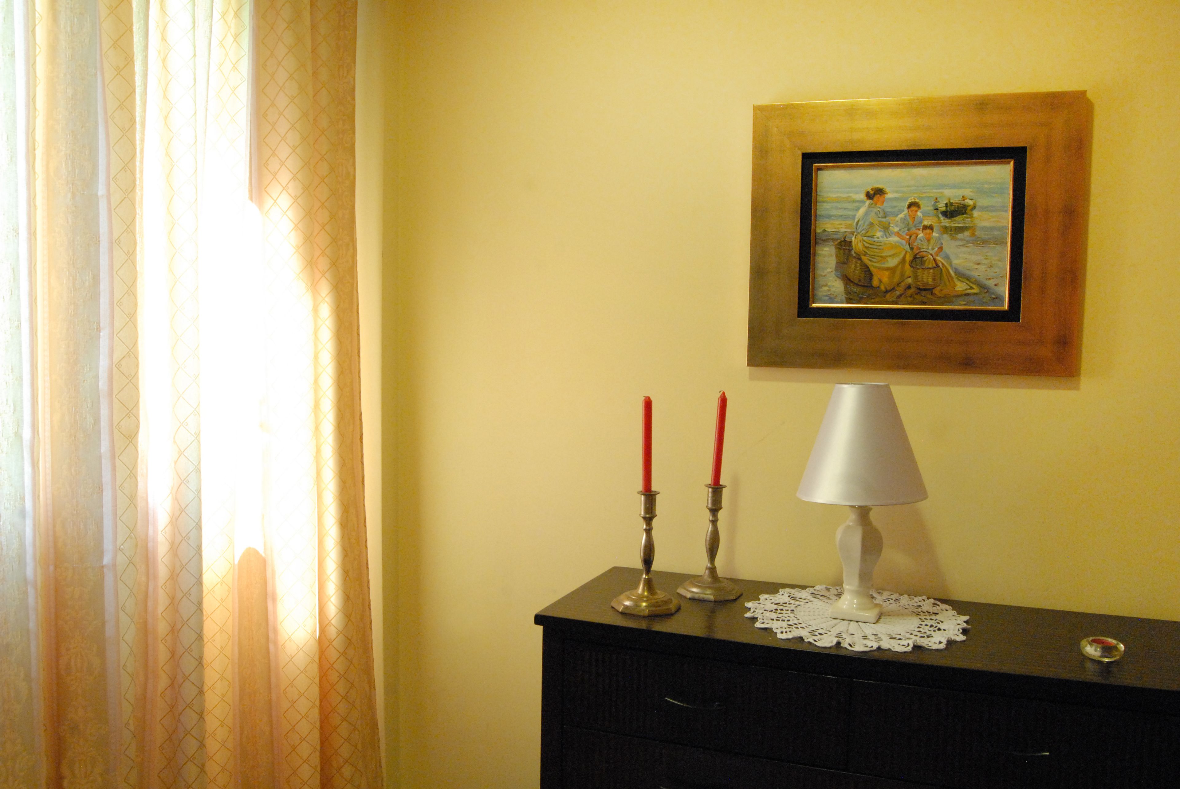 Una habitación con un aparador de madera oscura, un cuadro enmarcado, dos velas rojas y una lámpara de mesa blanca.
