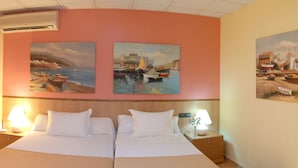 A hotel room with a bed, two bedside tables with lamps, and three paintings on the wall depicting coastal scenes.