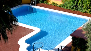 A rectangular swimming pool with clear blue water, surrounded by a red and yellow tiled deck, and bordered by lush green plants.