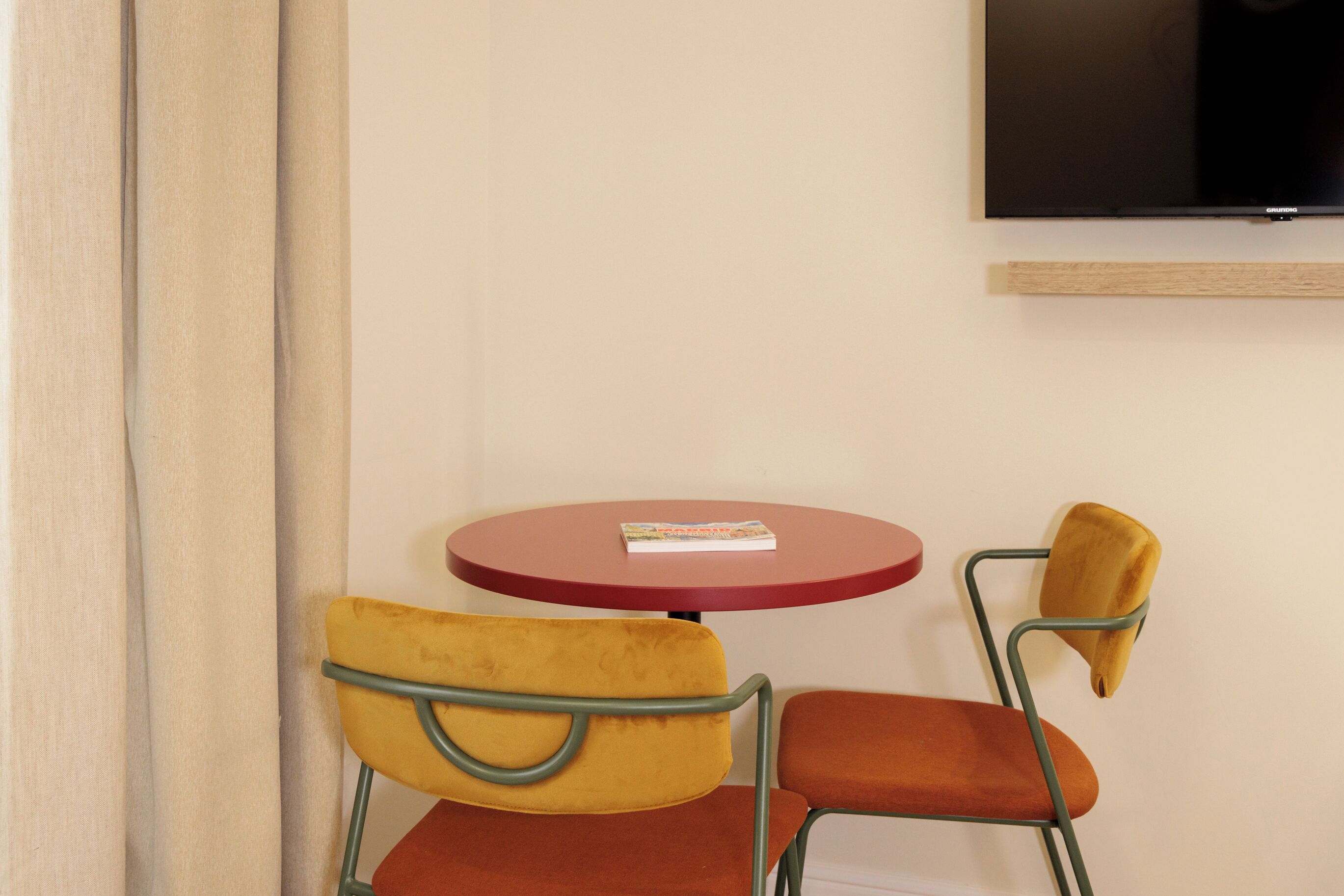 Een kamer met een aan de muur bevestigde televisie, een ronde rode tafel en twee stoelen met metalen frames en beklede zitplaatsen.