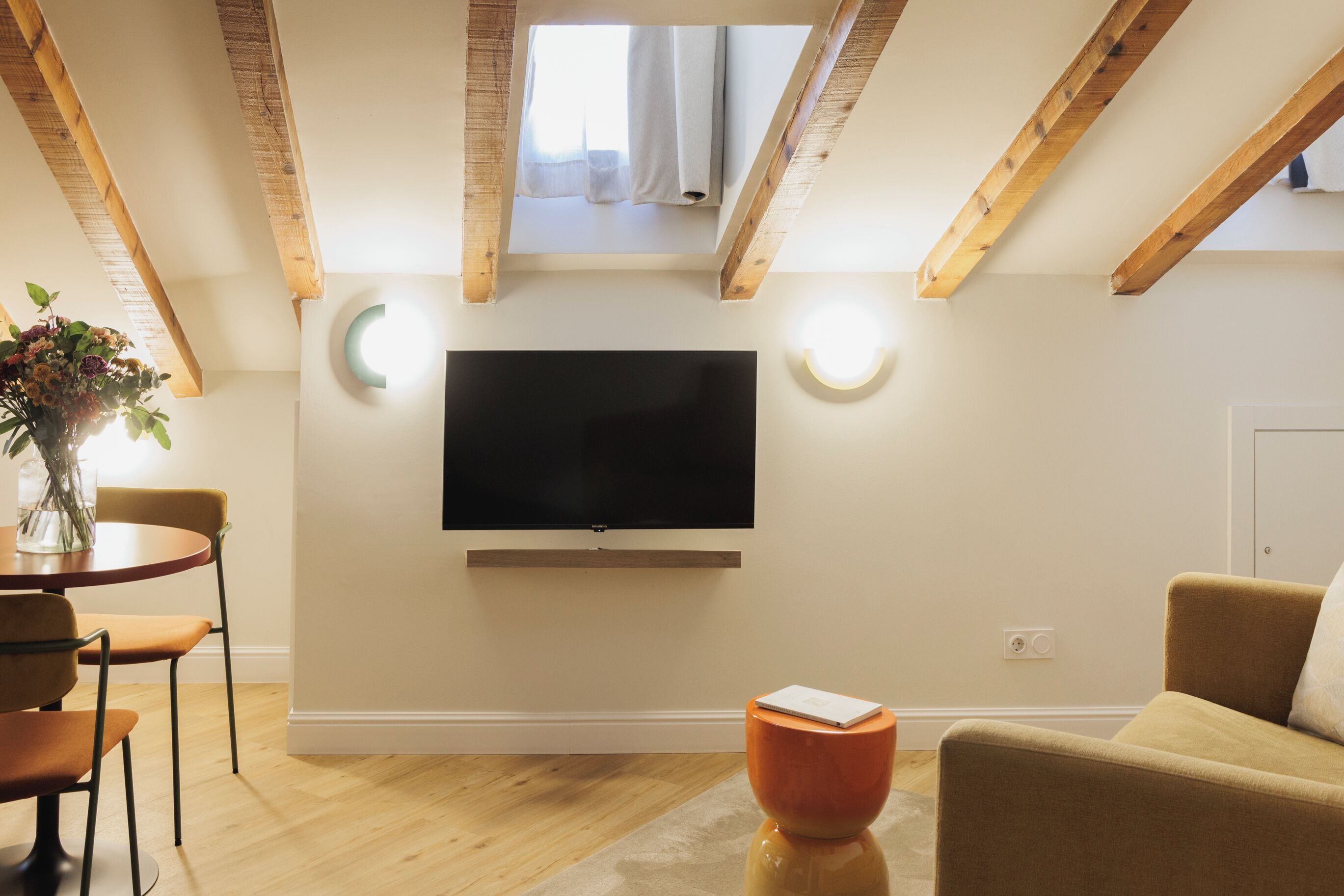 Een moderne woonkamer met een flatscreen televisie, houten balken, een eethoek met stoelen en een kleine tafel met een vaas.