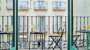 Un balcón con una silla de madera y una mesa, una ventana con persiana cerrada y una barandilla de metal.