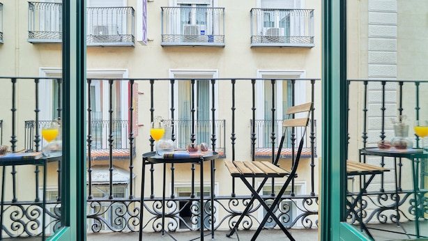 Un balcón con una silla de madera y una mesa, una ventana con persiana cerrada y una barandilla de metal.