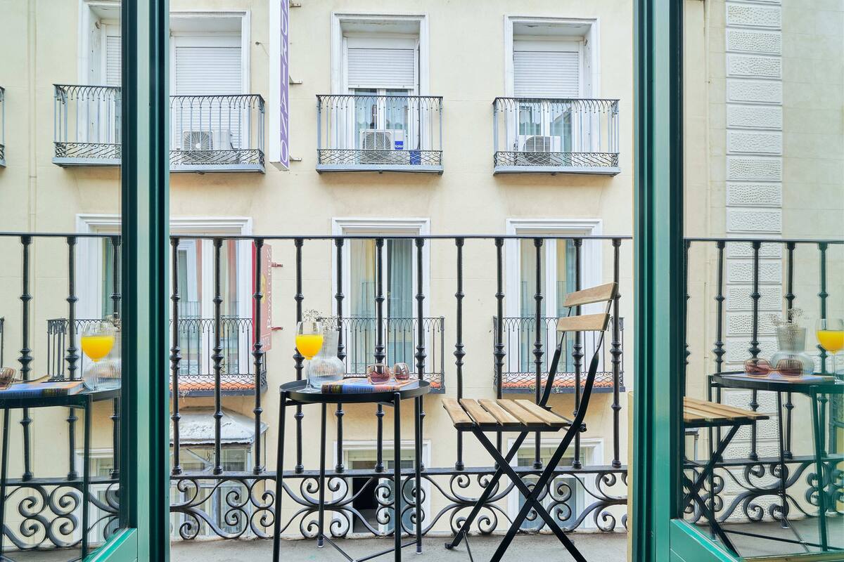 A balcony with a wooden chair and a table, a window with a closed shutter, and a metal railing.