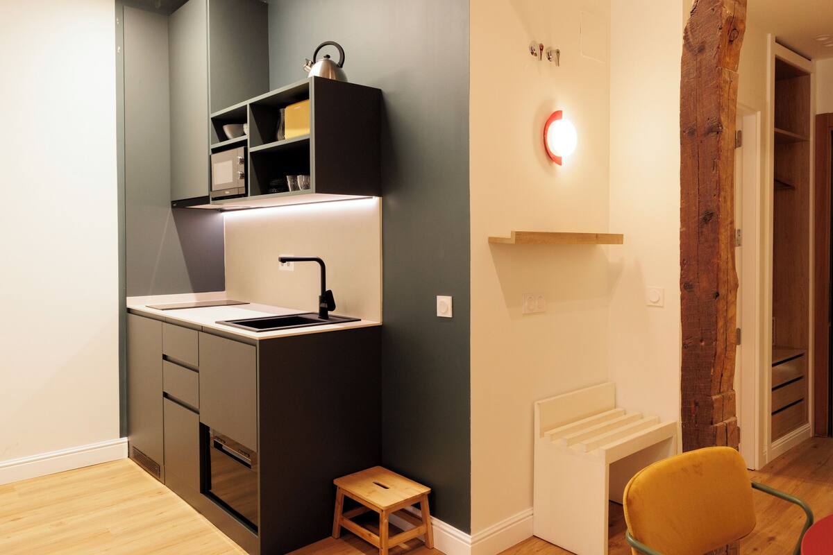 A compact kitchen with a sink, cabinets, and a stool.