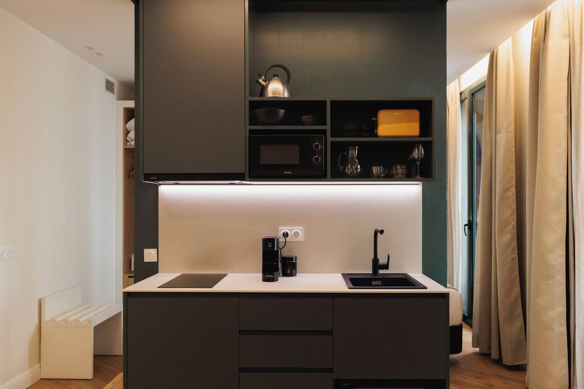 A compact kitchen with a microwave, kettle, and coffee maker, a small sink, and a cabinet with glassware.