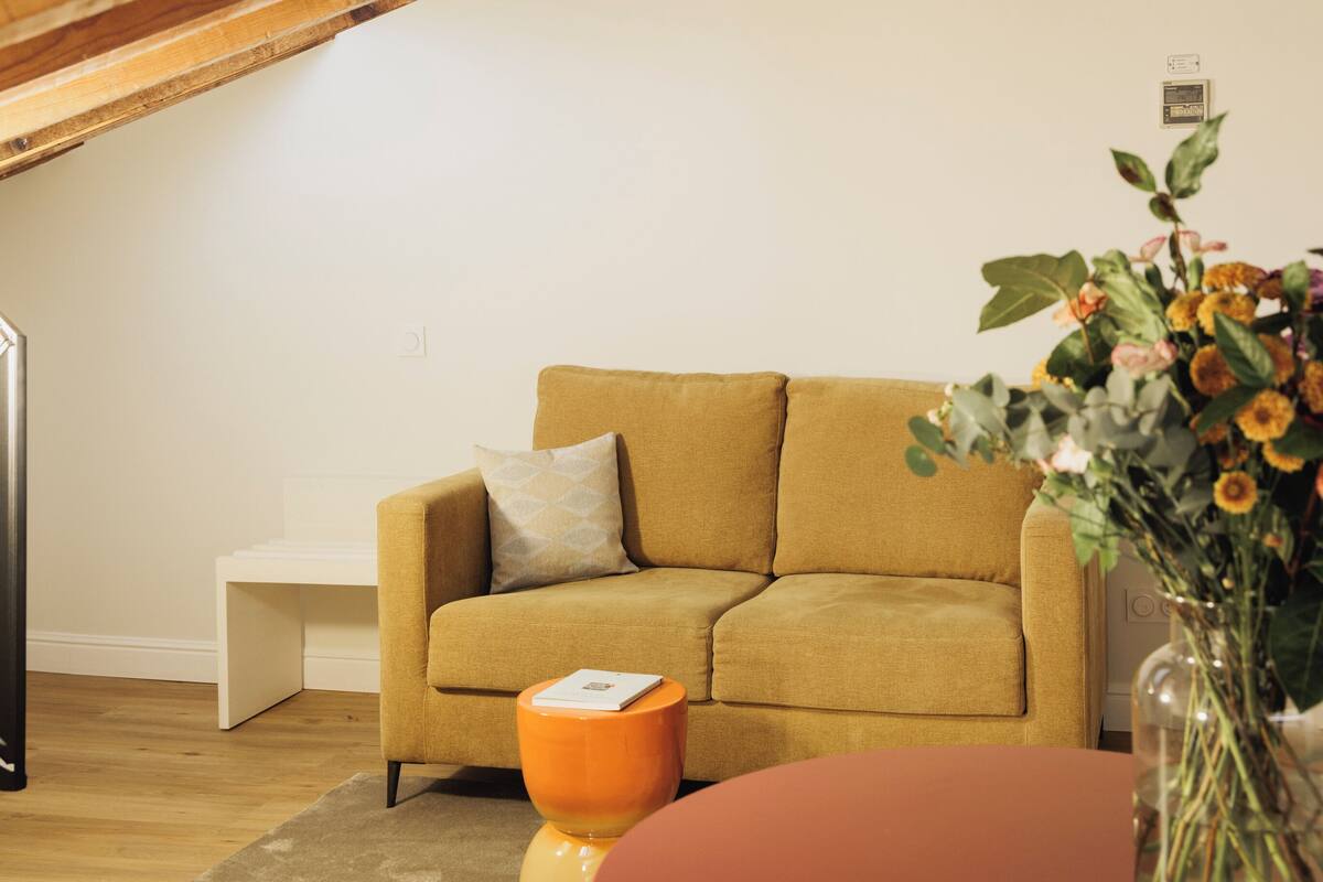 A modern living room with a sofa, a small orange side table, a vase with flowers, and a white wall.