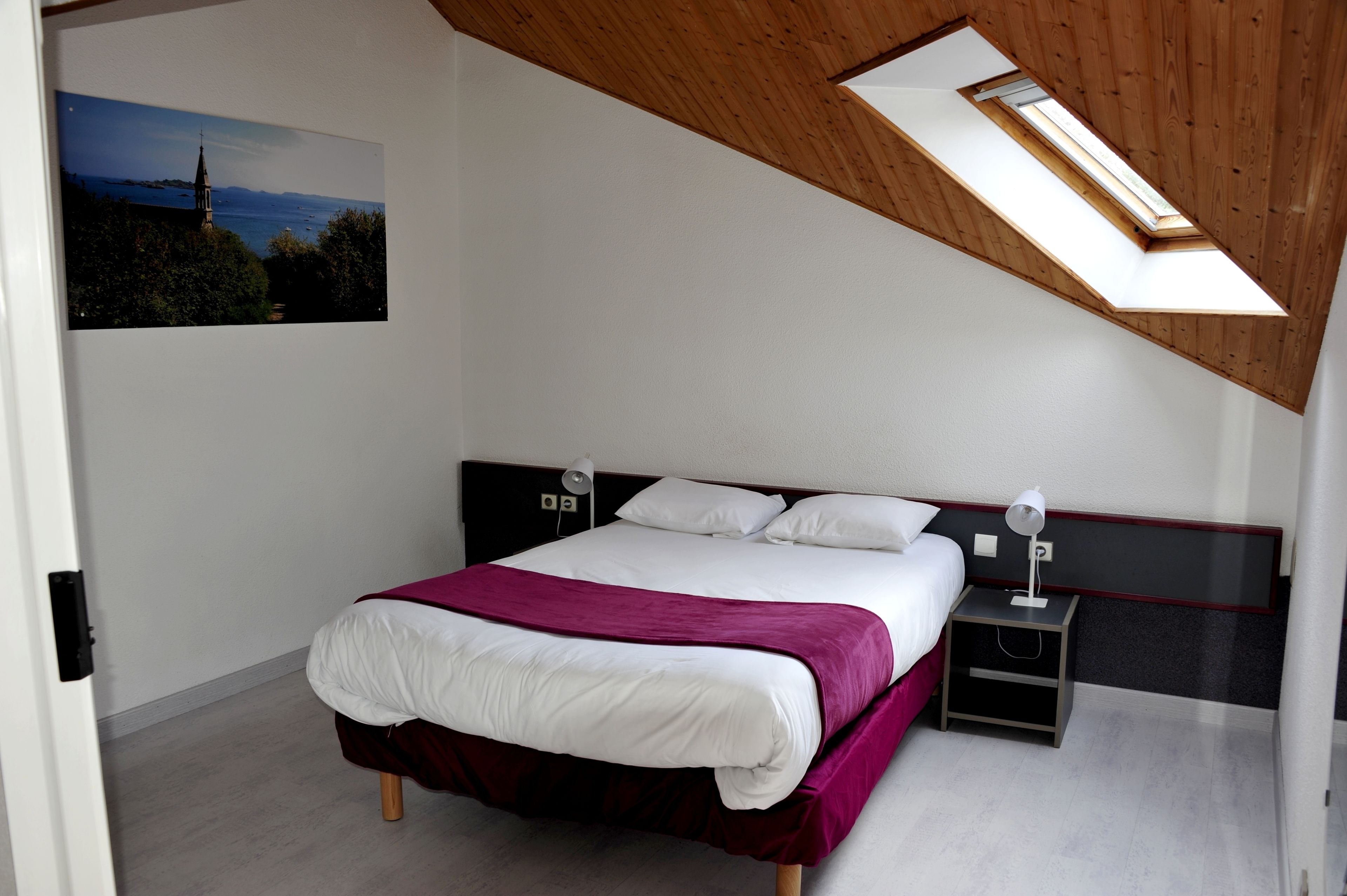 Un dormitorio con una cama, una mesita de noche, una lámpara de pared y un tragaluz.