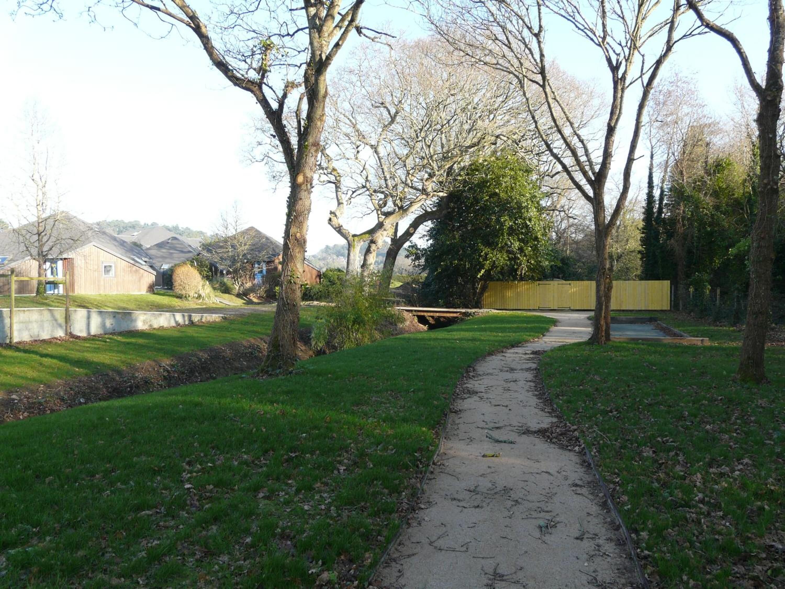 A pathway leading through a park with trees and a small bridge over a water body.