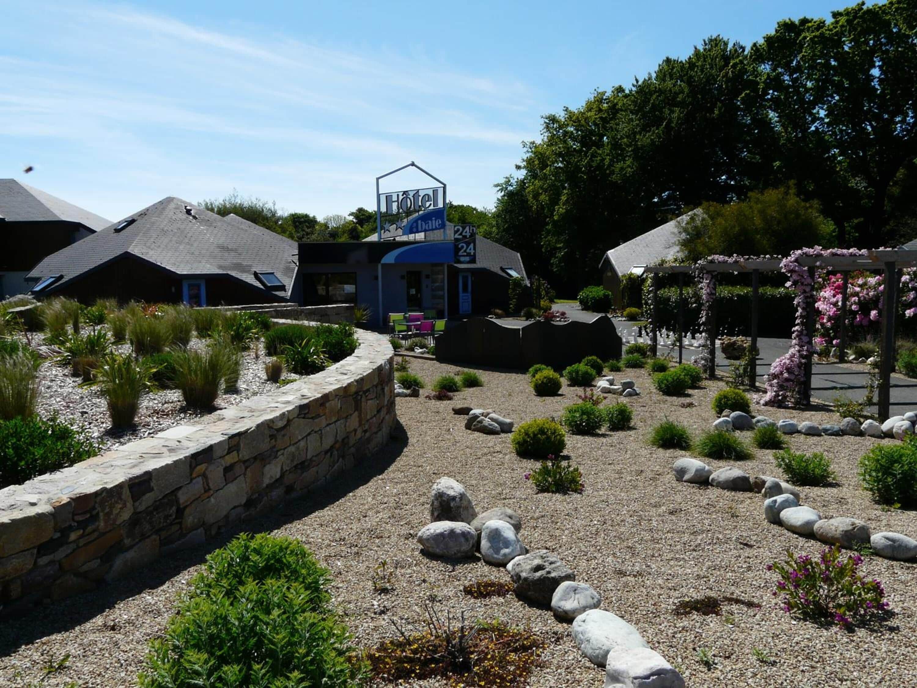 Un hotel con un muro de piedra, un jardín con diversas plantas y rocas, y una pérgola con flores rosadas.