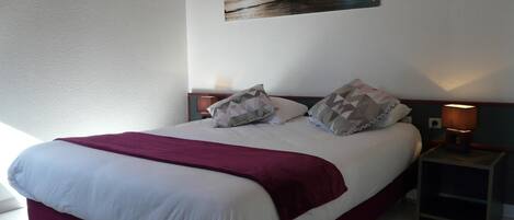 Una cama bien hecha con colcha blanca y burdeos, dos almohadas y una manta burdeos. Hay una mesita de noche con una lámpara y un pequeño estante. Sobre la cama, en la pared, cuelga un cuadro enmarcado con una imagen de un paisaje marino.