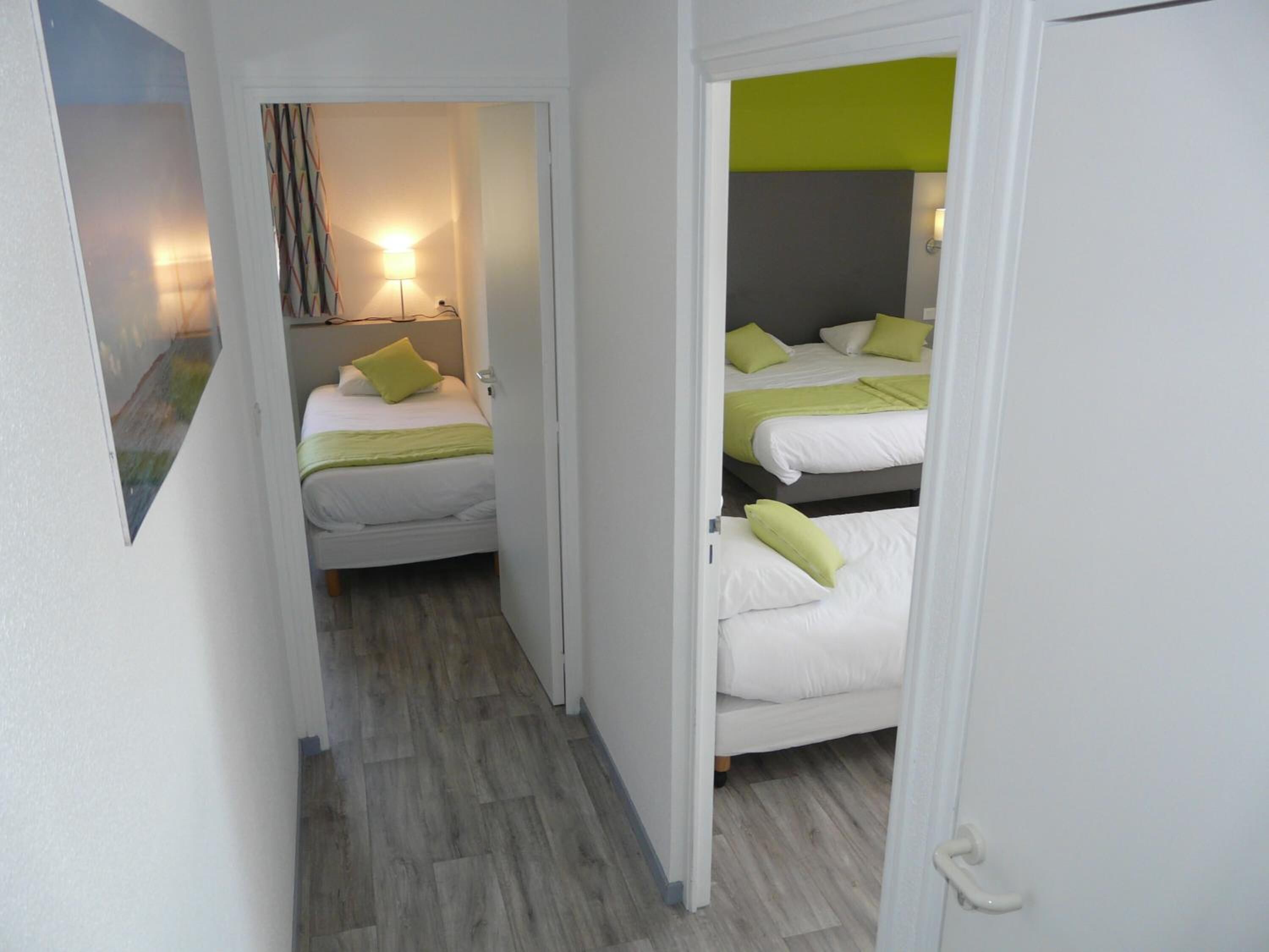 Ein schmaler Flur, der zu einem Zimmer mit zwei Betten führt, beide mit weißer Bettwäsche und grünen Kissen.