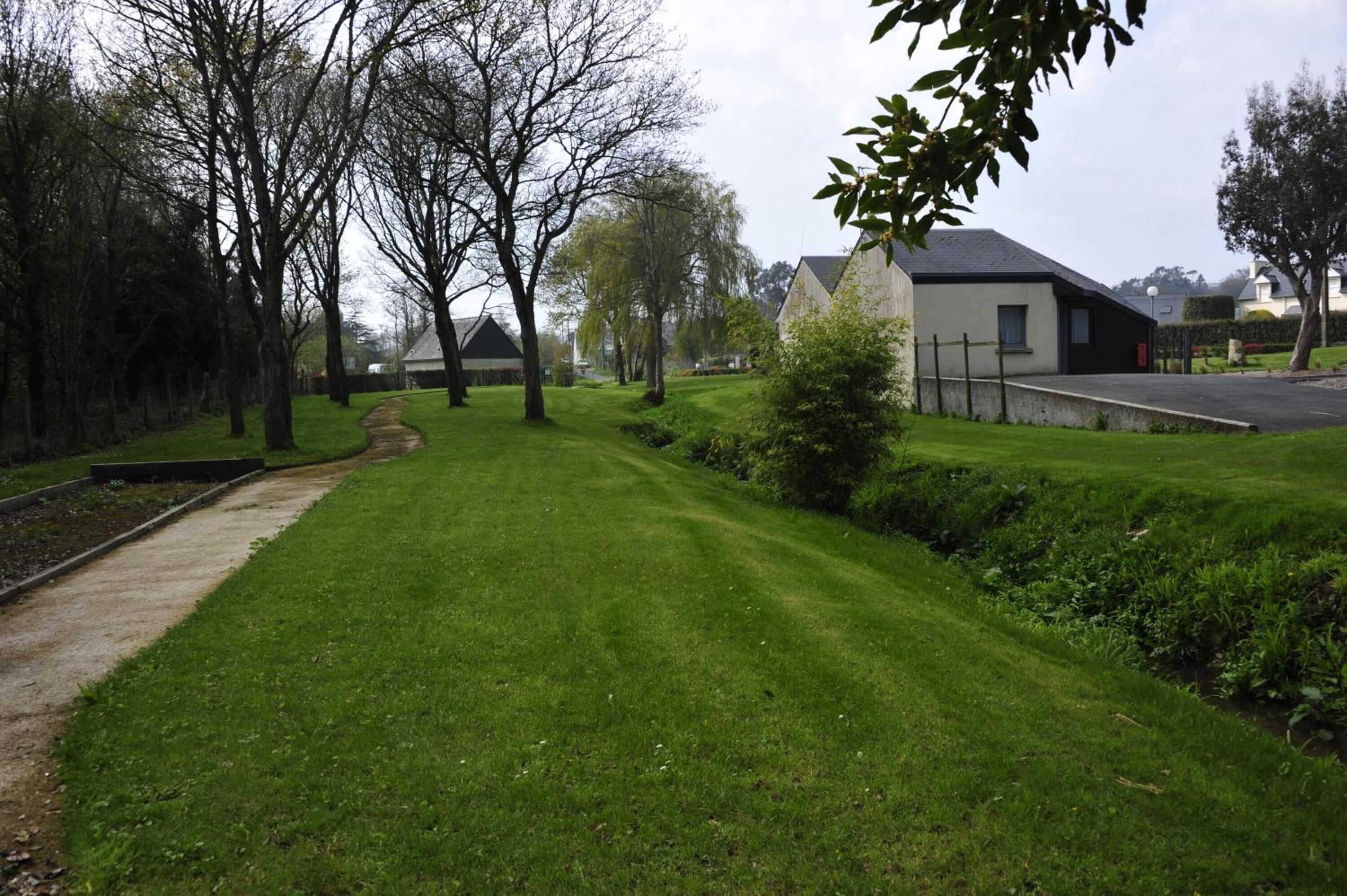 A well-maintained grassy area with a pathway, trees, and a building with a dark roof and white walls.