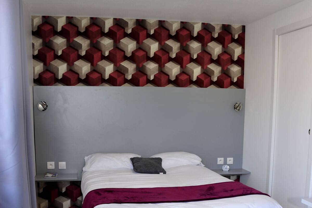 A neatly made bed with a white comforter and a maroon throw blanket. There are slippers and a folded towel on the floor.