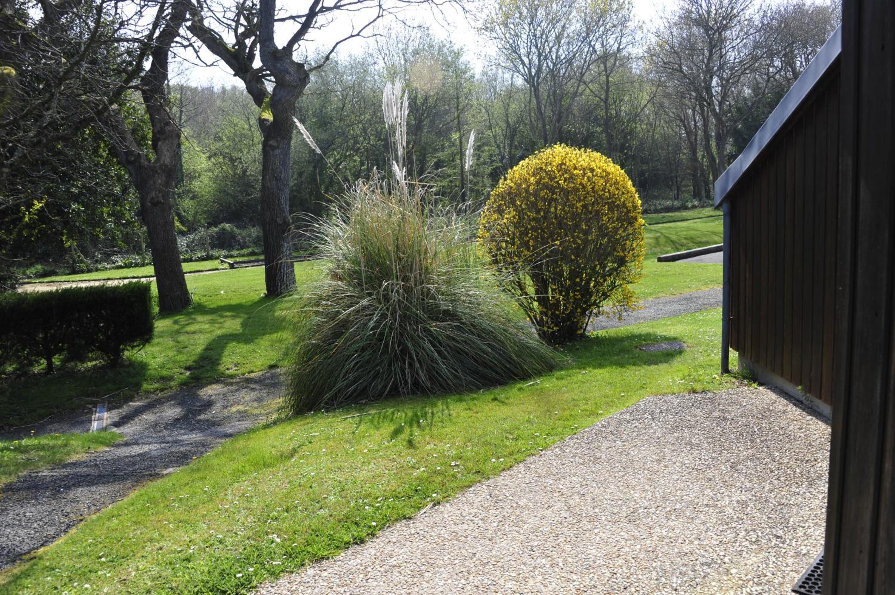 A gravel pathway leading to a wooden building, surrounded by manicured lawns and various shrubs.