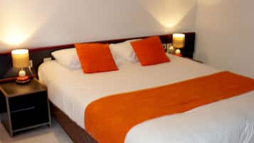 Una cama bien hecha con ropa de cama blanca y naranja, dos mesitas de noche con lámparas, y un cuadro con una imagen de un velero en la pared.