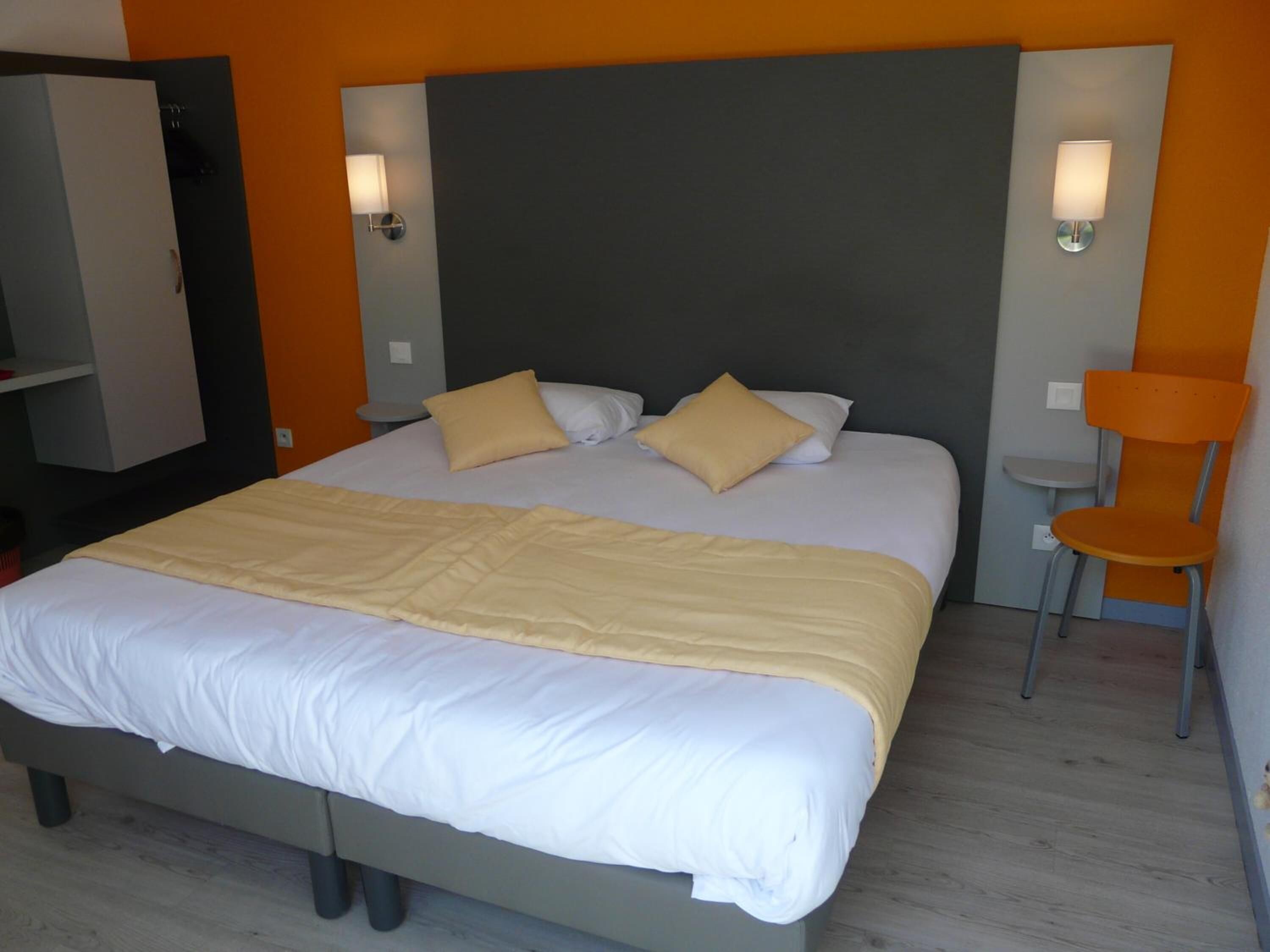 A modern hotel room with a large bed, two bedside lamps, an orange chair, and a wardrobe.