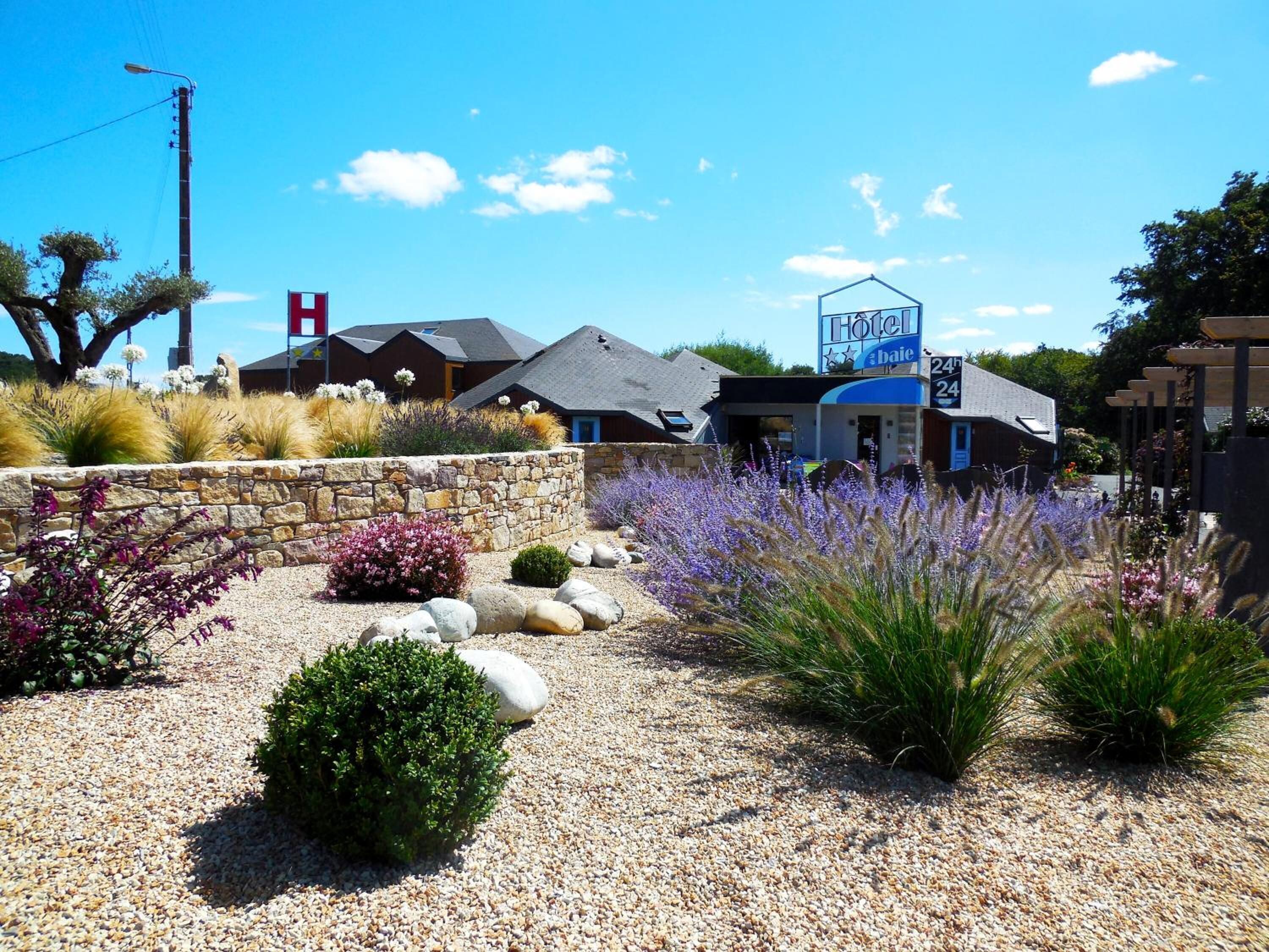 A hotel with a blue sign, stone wall, and gravel pathway with various plants.